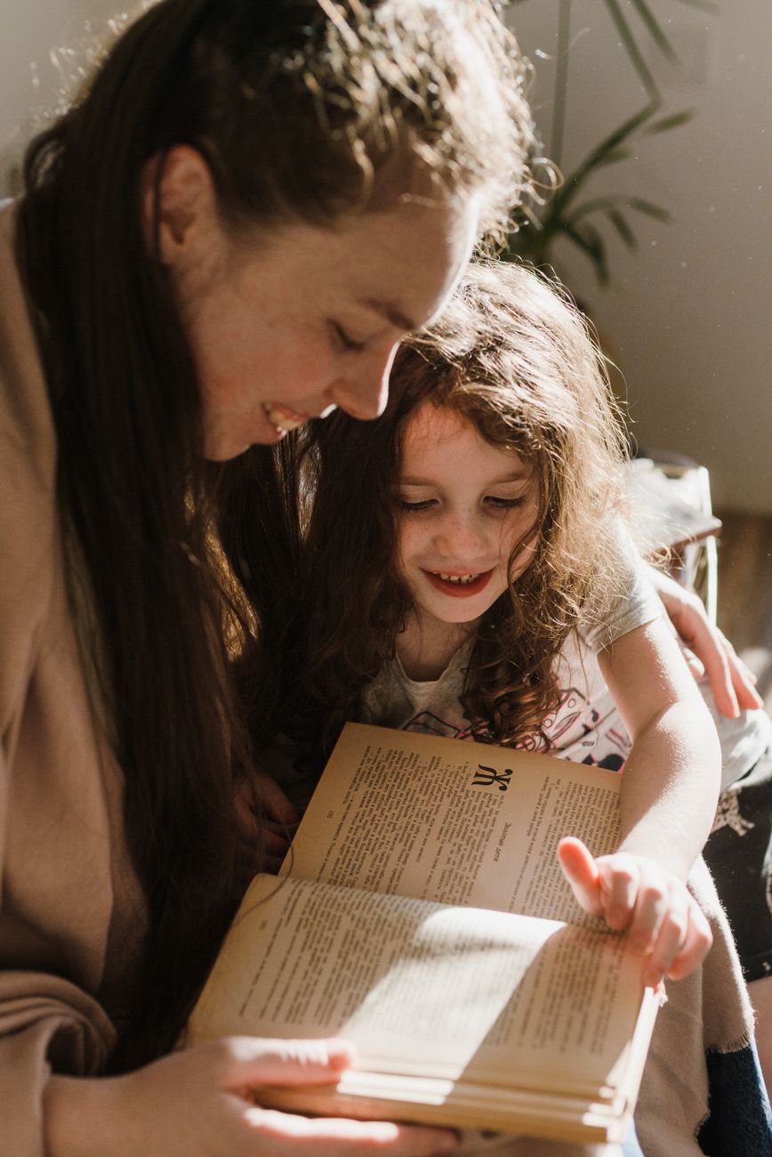 A mother and daughter share a joyful moment reading a book together in a warm, sunlit room.
